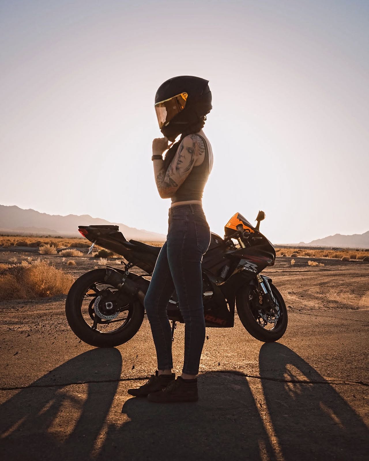 Rainey in a full-face helmet standing beside her Suzuki GSX-R in the desert at sunset